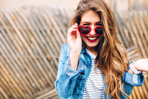 smiling woman in denim jacket and  red heart shaped sunglasses standing in front of wooden fence